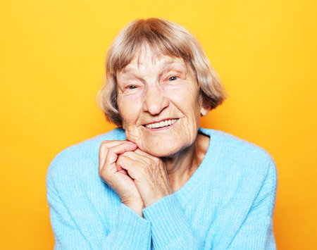 Emotion, lifestyle and old people concept: Happy elderly woman with wrinkles on face wearing blue sweater smiling at the camera over yellow backgoundの写真素材