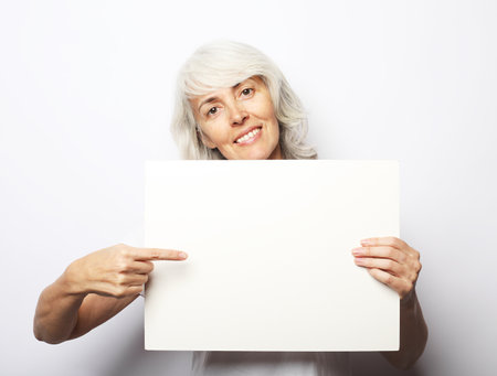A happy and charming elderly woman holds a blank sheet of paper in her hands and points at it with her finger. Sjot over white background.の写真素材