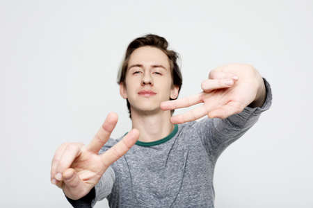 young man posing in white isolated studio background and showing the victory sign, looking at the camera, casual styleの写真素材