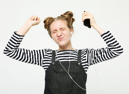Portrait of a cheerful stylish young woman wearing jeans sundress and striped shirt standing isolated over white background, listening to music with headphones, dancingの写真素材