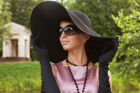 Beautiful elegant young lady enjoying a day in the park. Big black hat and sunglasses.の写真素材