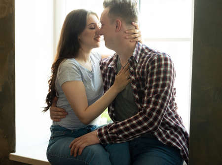 Young couple hugging and kissing while sitting on the windowsill by the window