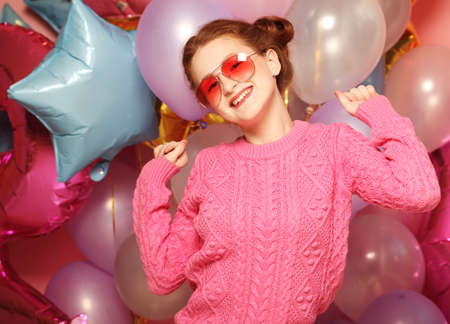 Funny young woman in pink sweater isolated on bright balloons background.の写真素材