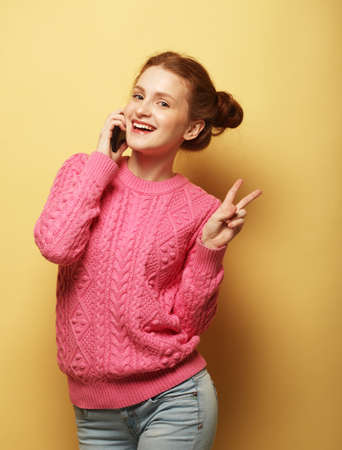 Young redhead woman wearing pink sweater with smart phone show v-sign over yellow backgroundの写真素材