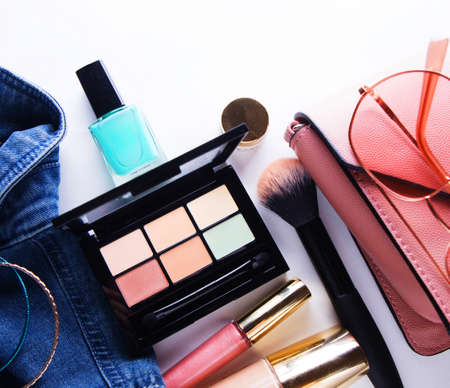 Top view of woman jeans shirt, concealer pallet, lip gloss, nail polish, bracelet, powder brush on...
