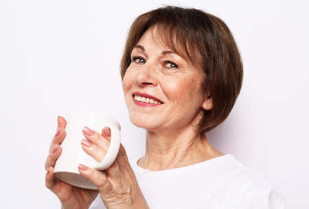 Old excited lady smiling laughing, holding cup drinking coffee, tea, beverage on white backgroundの写真素材