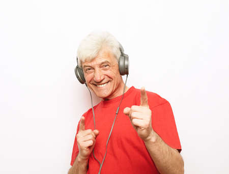 Handsome old man in casual wear and headphones is listening to music and show ok sign on white...