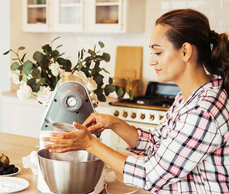 Young woman uses mixer and whips cream for cake, stands in modern kitchen