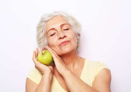 Portrait of a beautiful elderly woman with closed eyes holding an apple, smiling, isolated on white...