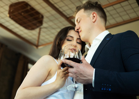 The groom in a suit and the bride in a wedding dress hold glasses of red wine drinking.の写真素材