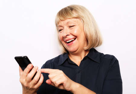 Pretty senior female in casual outfit using modern smartphone and laughing out loud while standing on white background. Old woman watching funny videos on internet.の写真素材