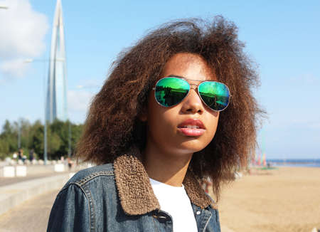 Young african woman in a denim jacket and sunglasses is resting while sitting on the beach on a...
