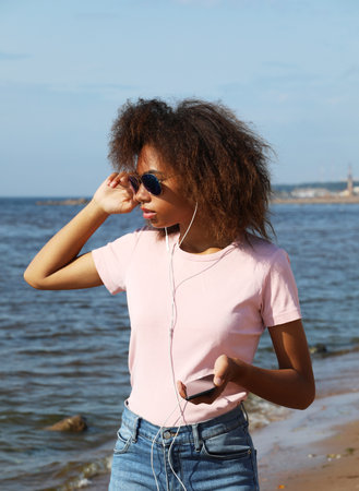 Young afro american woman in sunglasses listening to music in headphones on her mobile phone near sea.の写真素材