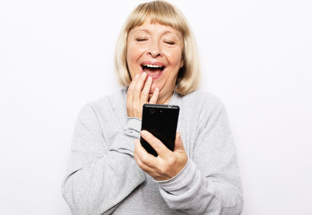 Image of a happy surprised shocked old woman posing isolated over white background using mobile phone.の写真素材