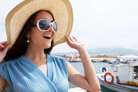 Beautiful young brunette woman in blue dress and hat stands at the port and looks at the boats, summer time.の写真素材