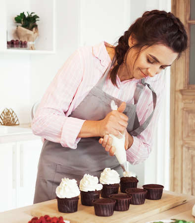 Young woman with confectionery bag squeezing cream on cupcakesの写真素材