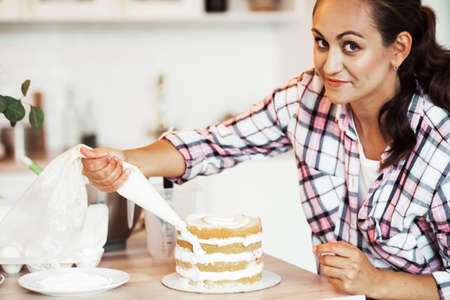 Beautiful pleased pastry chef woman making cake with cream at cozy kitchenの写真素材