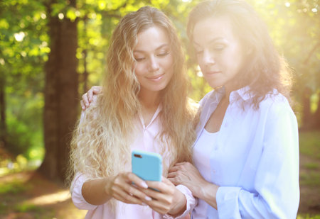 Portrait of Adult caucasian daughter showing on mobile phone to her senior mother outdoor in natureの写真素材
