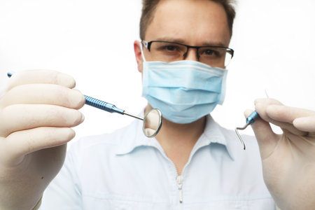 Young dentist male in white lab coat and mask holding dental instruments, isolated on white backgroundの写真素材