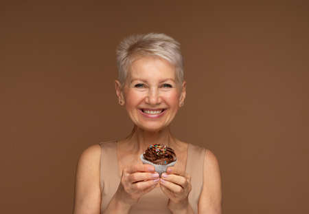 Elderly woman with short hair holds a chocolate cupcake in her hands.