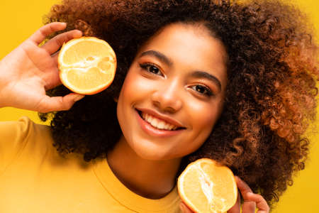 Healthy summer, diet and people concept: Cheerful afro woman holding oranges near ears, yellow...