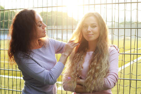 Mother straightens her adult daughter's hair, sunset time, park. lifestyle.の写真素材