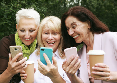 Three elderly women are smiling and looking photos at the at screen of the phones in the park on a...