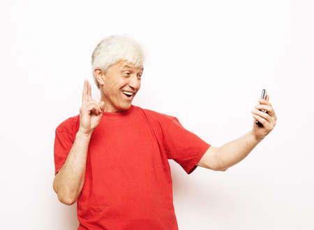 Modern lifestyle, technology and old people concept: An elderly man dressed in a red t-shirt holds a smartphone and takes a selfie. Pensioner smiles and rejoices while standing on a white background..の写真素材