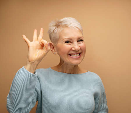 Happy smiling old lady with short hair showing ok sign headshot at studio. Elderly woman giving good...