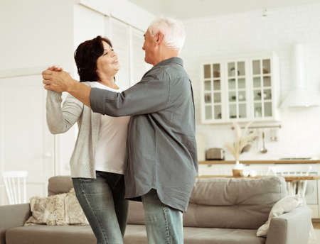 Carefree happy active old senior couple dancing in kitchen, cheerful retired elder husband holding...
