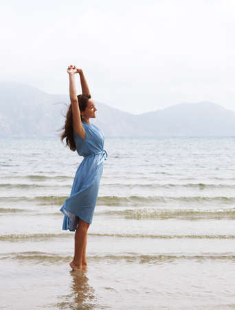 Young brunette woman in a blue dress walking barefoot on a beach and dangles his feet in the water. Happy summer holiday.の写真素材