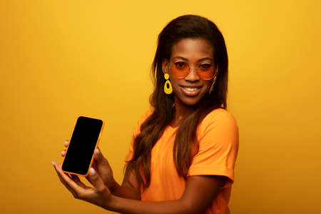 Young smiling african american woman isolated on yellow background, wearing orange t-shirt, holding blank phone and showing interesting offer over yellow backgroundの写真素材