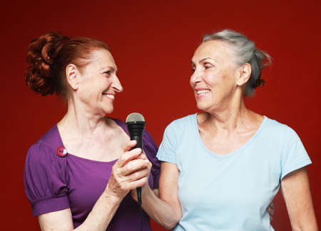 two elderly female friends with microphone, laughs and prepares for party karaoke over red backgroundの写真素材