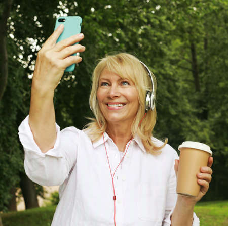 Beautiful elderly pensioner woman in headphones holding coffee to go and smartphone.