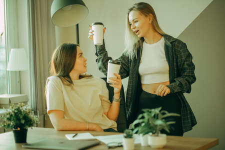 Two young female colleagues drink coffee in the office and discuss work.