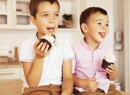Two brothers male kids are sitting together at the kitchen table and eating cupcakesの写真素材