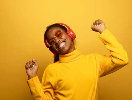 People, music, emotions concept. Young afro american female with dances in rhythm of melody, closes eyes listens loud song in headphones over yellow background.の写真素材