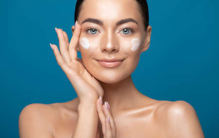 Beautiful young woman applying cosmetic cream treatment on her face isolated on blue color backgroundの写真素材
