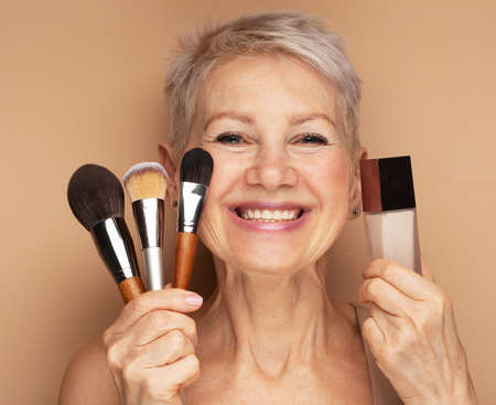 Charming elderly woman holds makeup brushes and foundation in her hands. beauty at any age.の写真素材