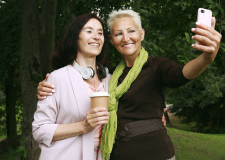 Two charming elderly women friends holds a cup of coffee to go and making selfie in summer park.の写真素材