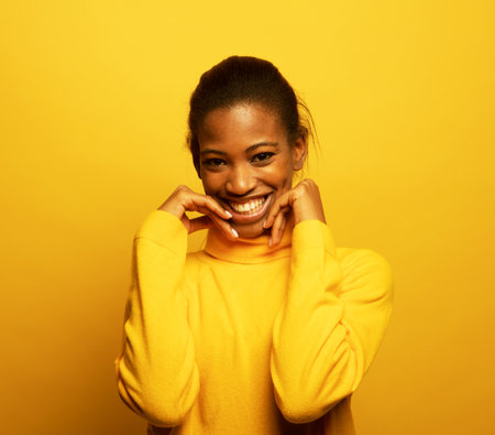 Charming afro american woman in yellow sweater smiles happily pressing her hands to her face. Appreciation and trust, happiness concept. Portrait on a yellow studio background.の写真素材