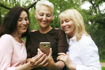 Three old happy women are smiling and looking photos at the at screen of the phone in the park on a summer dayの写真素材