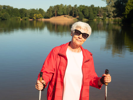 Gray-haired woman in sunglasses walking with tracking sticks on the beach near lake. Sport, healthy lifestyle and happy old age concept.の写真素材