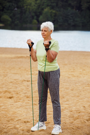 Old woman with short hair wear sportswear doing exercises outdoor on the beach in the morning using resistance rubber bands. Healthy lifestyle, active retired life and sporty time.の写真素材