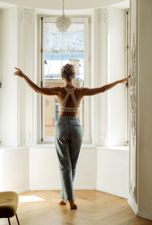A beautiful slim woman in jeans and a bra stands with her back to the camera and looks out the window, full length portraitの写真素材