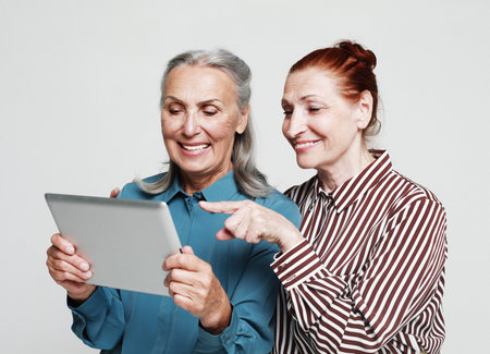 Lifestyle, friendship and old people concept. Two mature female friends looking through old photos digital album together with tablet device over gray background. Old friends Spending Quality Time Together.の写真素材