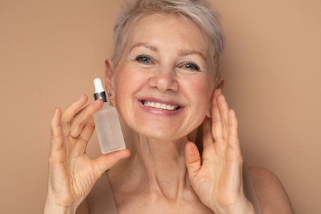 Happy smiling elderly attractive woman with gray hair holding in hand bottle with serum essence hyaluronic hydrate oil for skincare and haircare.の写真素材