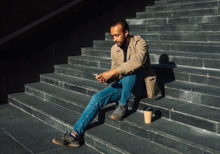 Portrait of young african american man wearing sunglasses sitting on steps using mobileの写真素材