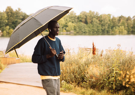 A young African-American man dressed in casual clothes walks on a cloudy morning in the rain next to a lake. The man is holding an umbrella. lifestyle concept.の写真素材