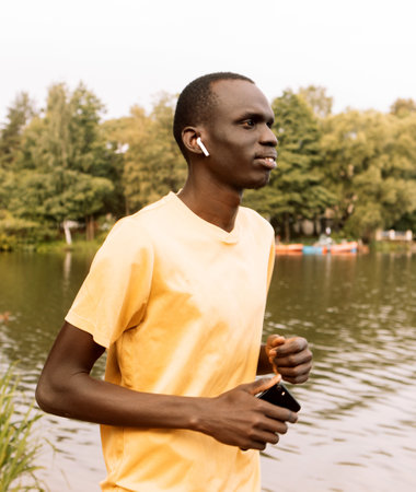 Cheerful black man wearing yellow t-shirt jogging outdoor along river bank early in the morning....
